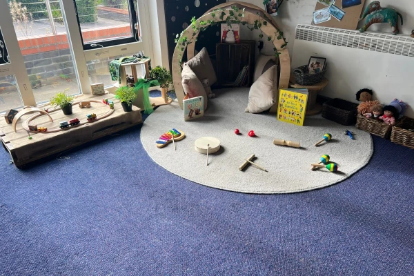 Semicircular wooden reading nook houses cushions and books; toys and wooden instruments lie scattered on a round rug, while a train set rests on a low table by windows.