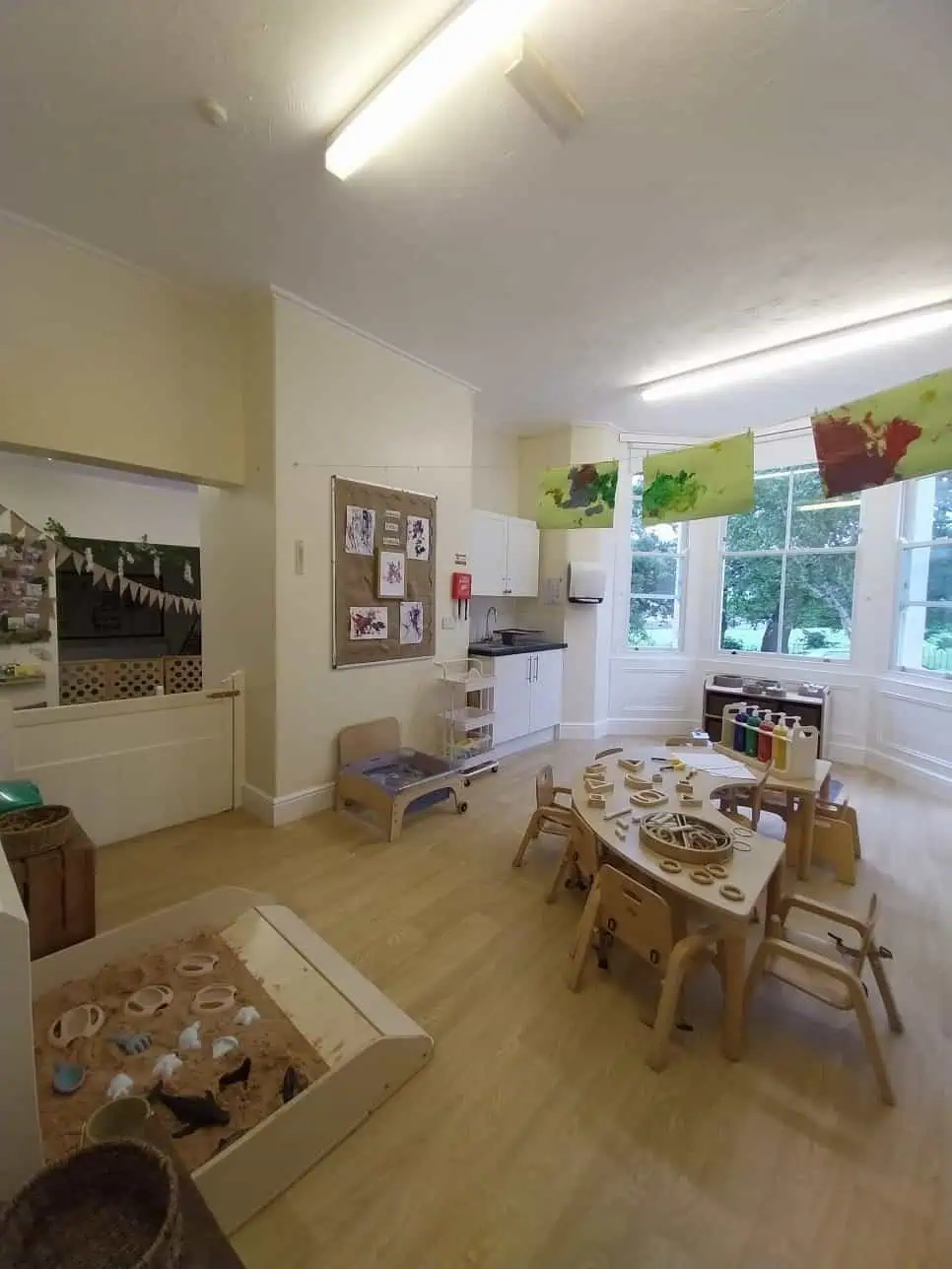 Low wooden table arranged with wooden rings and craft materials, surrounded by small chairs; artwork hangs overhead and a sand tray, sink, and large windows brighten the classroom.