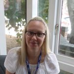 A seated woman with glasses and long blonde hair smiles while wearing a blue lanyard labeled 