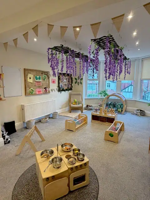 Wooden toy kitchen sits foreground, surrounded by low activity stations; artificial purple wisteria hangs above; bunting and large windows fill a bright, child-friendly classroom.