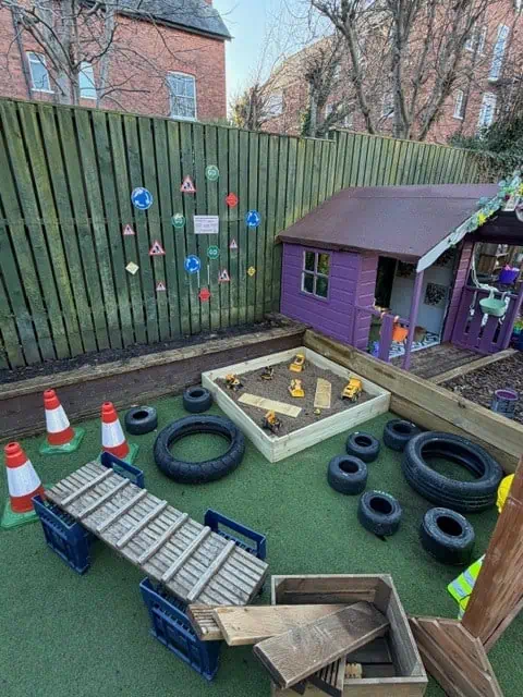 Purple wooden playhouse sits beside a sandbox with toy dump trucks; wooden ramps and plastic crates form bridges over artificial grass; traffic cones, rubber tires and road-sign stickers line a wooden fence.