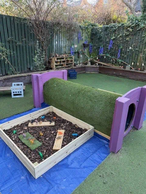 Purple play tunnel covered with turf sits on artificial grass; a wooden sensory box of mulch and toy animals sits beside it in an enclosed backyard with a green fence and climbing plants.