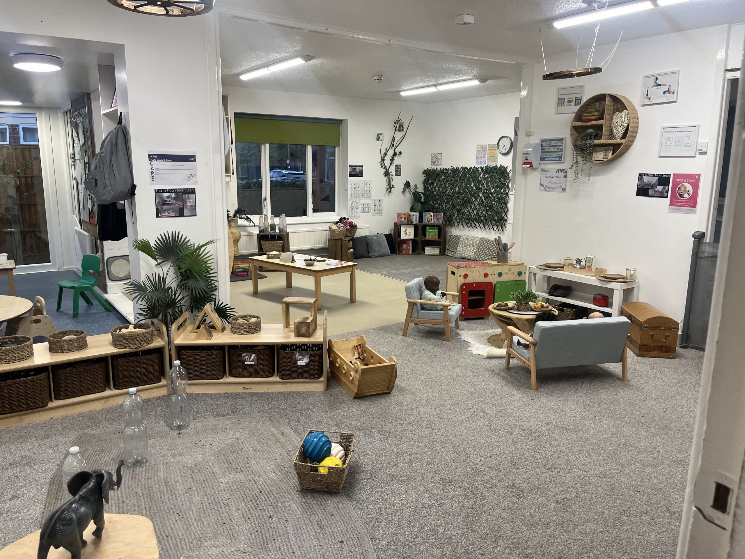 Children's play area — toys and small chairs arranged for pretend play in a carpeted nursery room with low tables, shelves, plants, and posters on the walls.