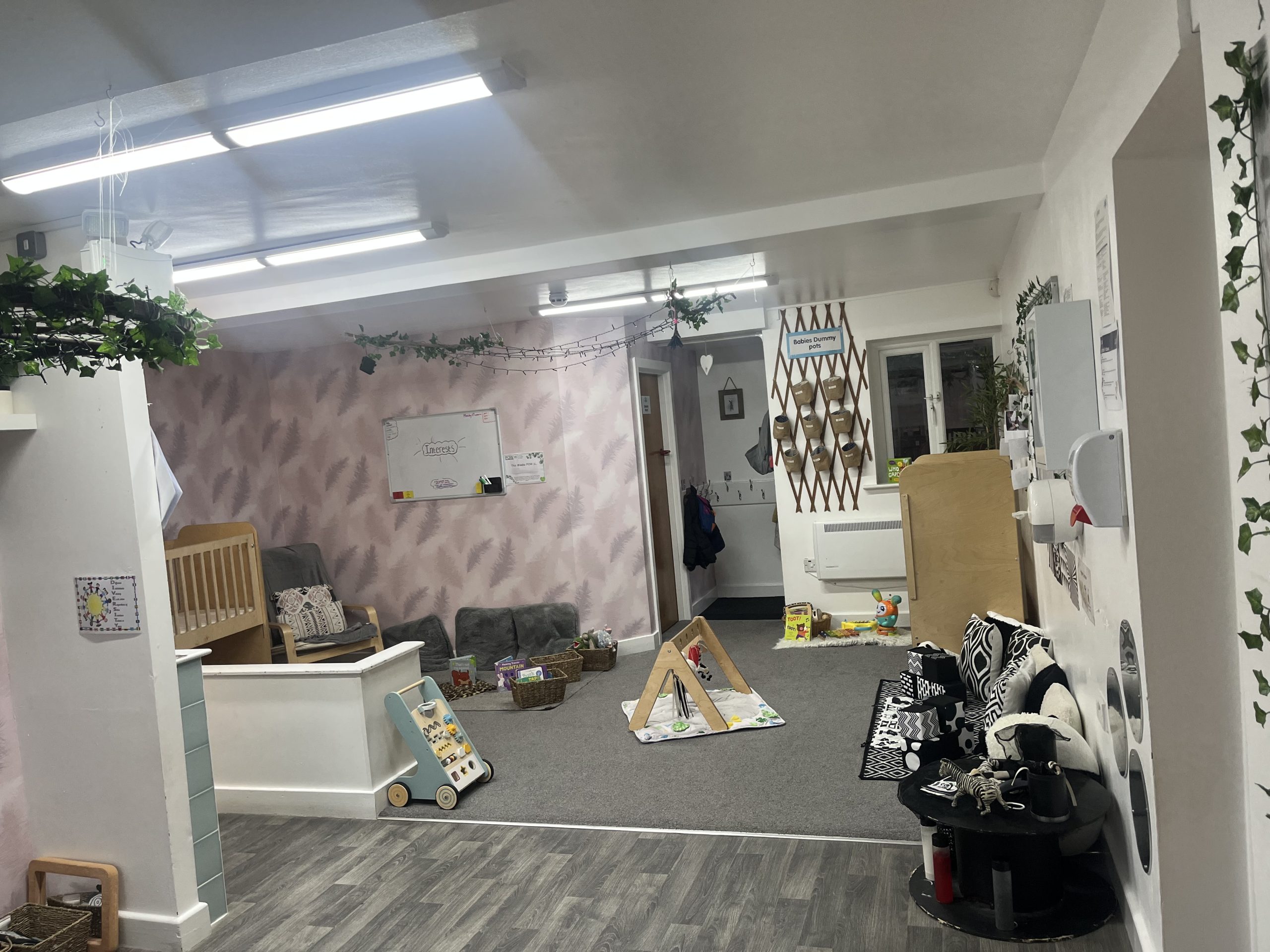 Small wooden baby gym sits on a play mat while toys, baskets and cushions are arranged around a carpeted nursery room beneath fluorescent ceiling lights.