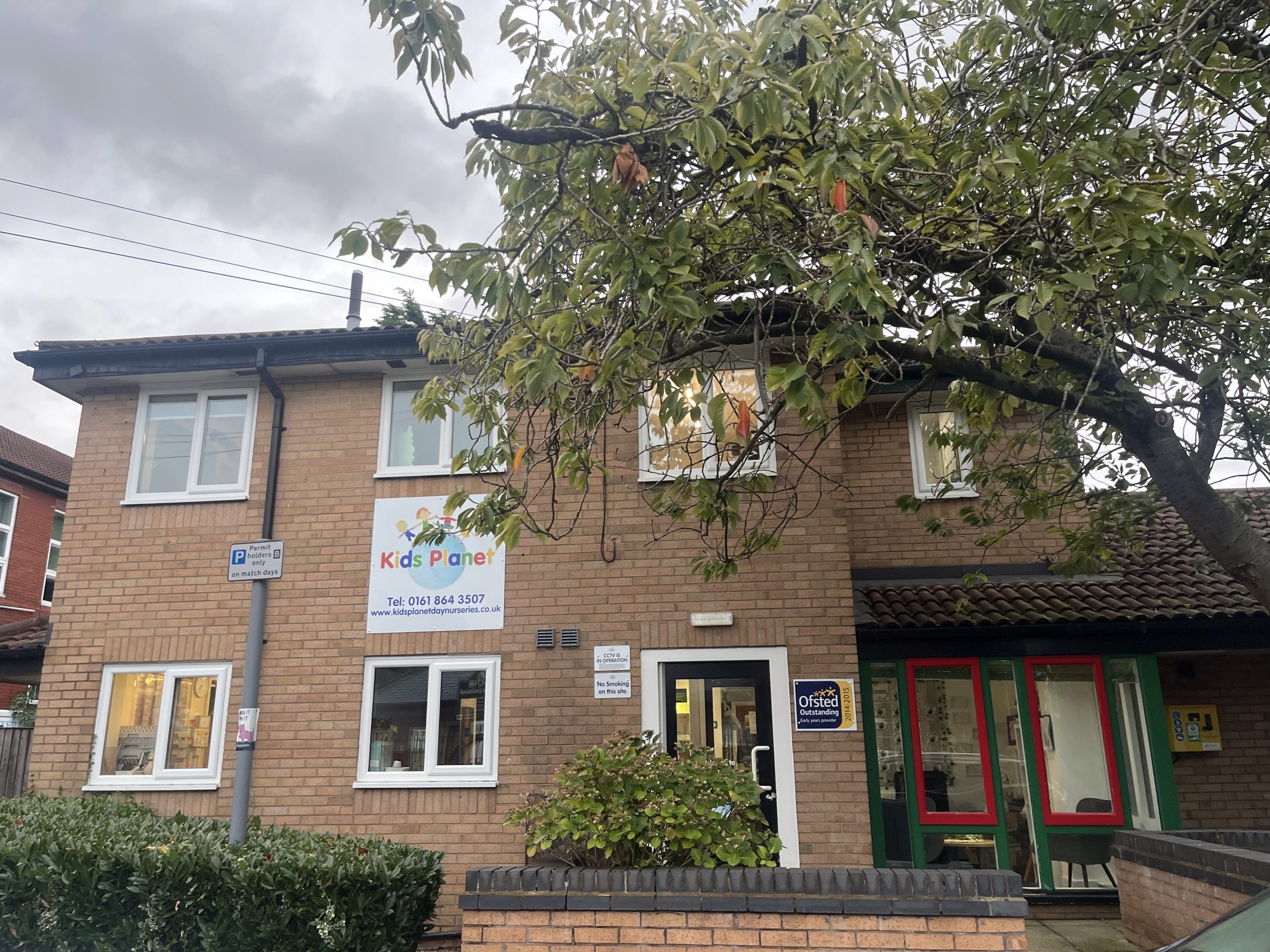 Kids Planet sign marks a two-story brick nursery; a tree overhangs the front entrance and windows, with hedges and a cloudy sky in the background.