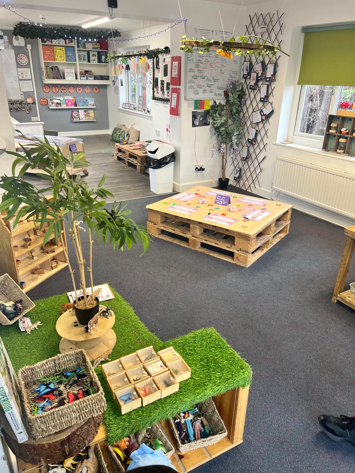 Low pallet activity table displaying paper name cards and small toys, surrounded by baskets of figurines, shelving, plants, and cushions in a bright early-years classroom/playroom.