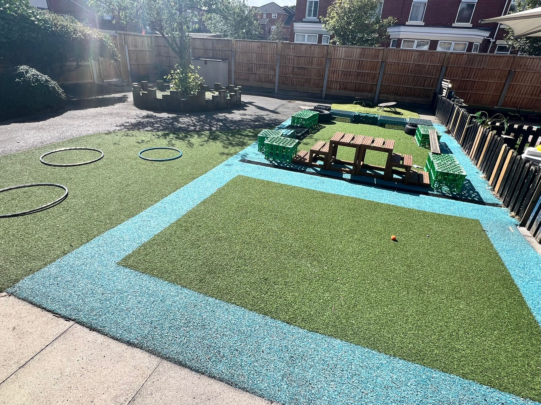 Artificial turf play area arranged with blue-bordered green mats, scattered hula-hoops, stacked green crates and small wooden benches, surrounding a tree planter inside a sunlit fenced backyard playground.