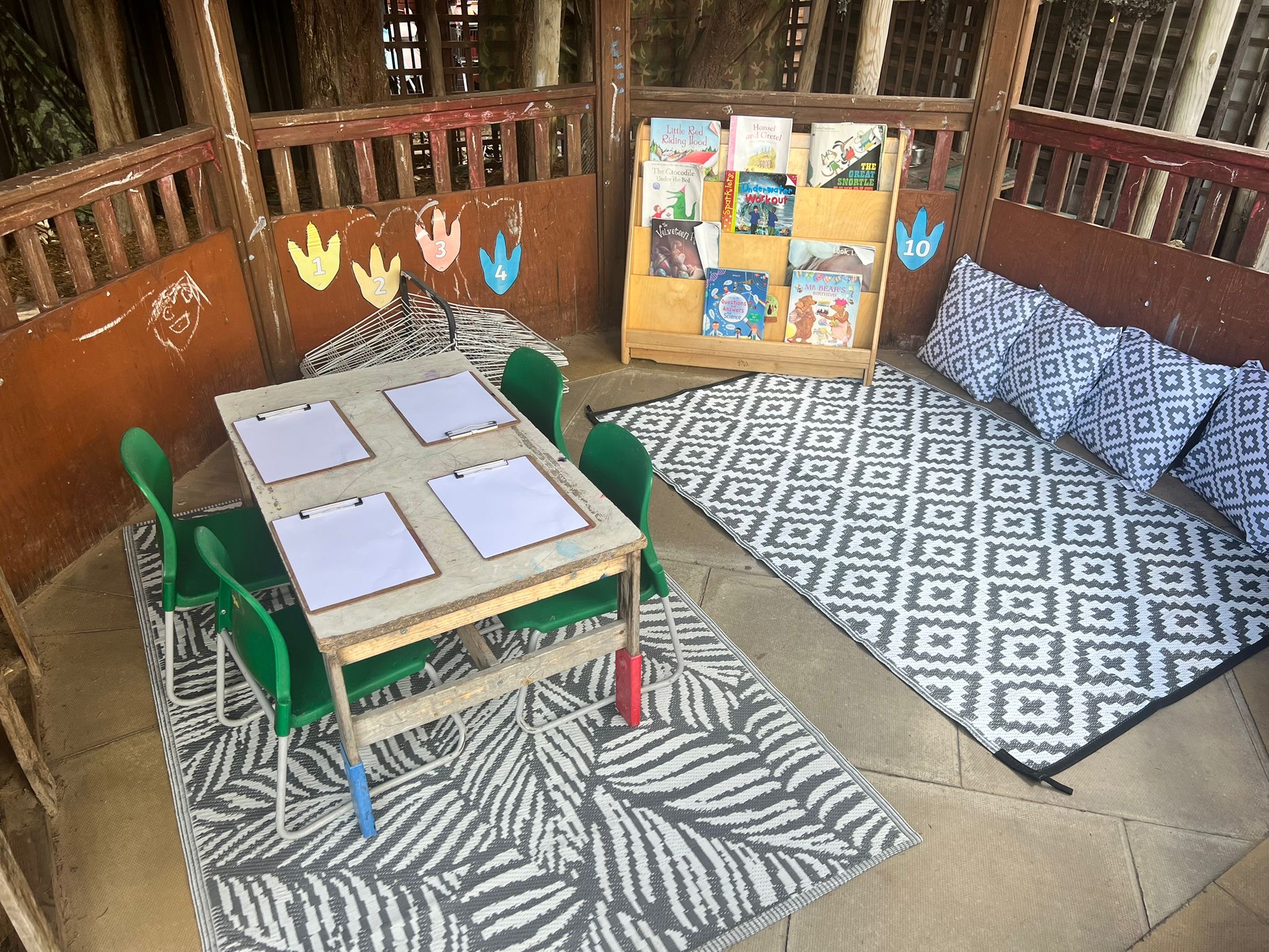 Small children's table with four clipboards, surrounded by green chairs; set inside a wooden playhouse with patterned rugs, cushions, a low book rack and numbered handprint labels.