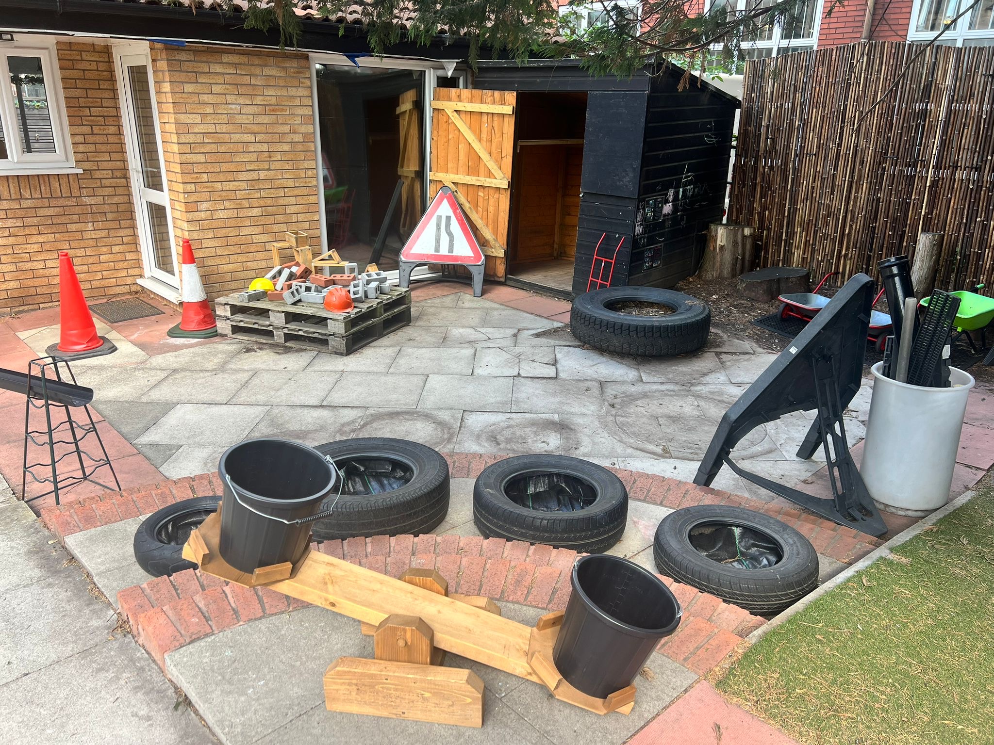 Wooden seesaw holding two empty black buckets sits atop a low brick edge, surrounded by stacked tires, traffic cones and construction blocks outside an open wooden shed in a backyard.