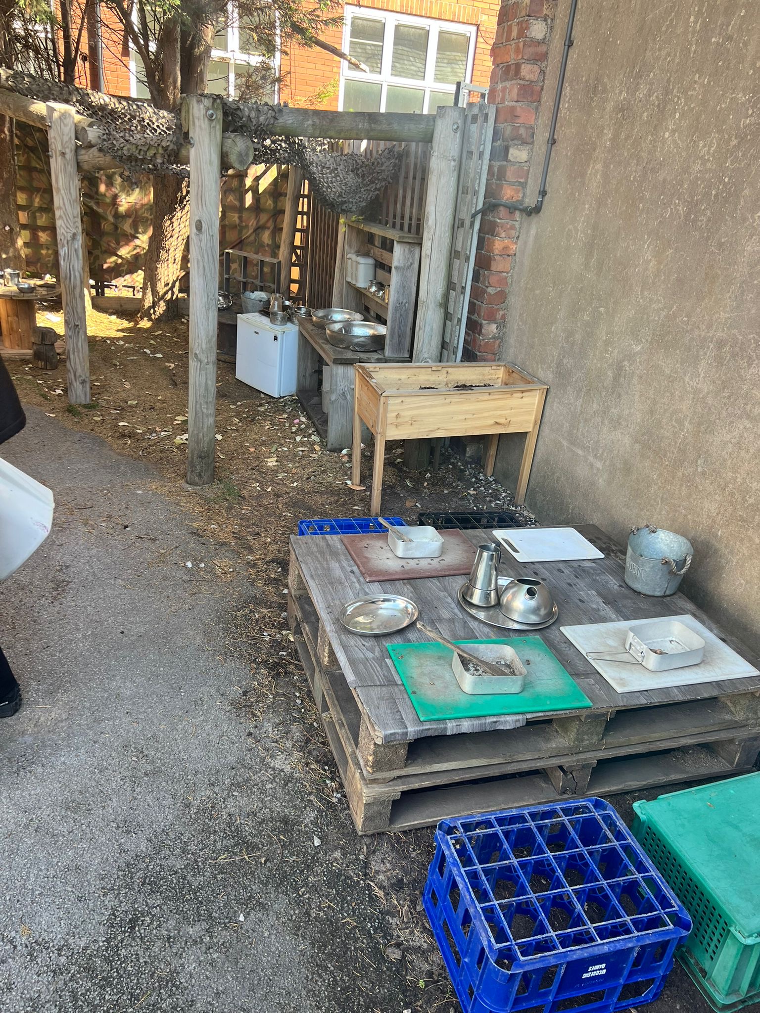 Wooden pallet table holds metal bowls, trays and kettles; blue crates nearby; a raised planter and rustic play kitchen sit beneath a pergola beside a brick wall in a yard.