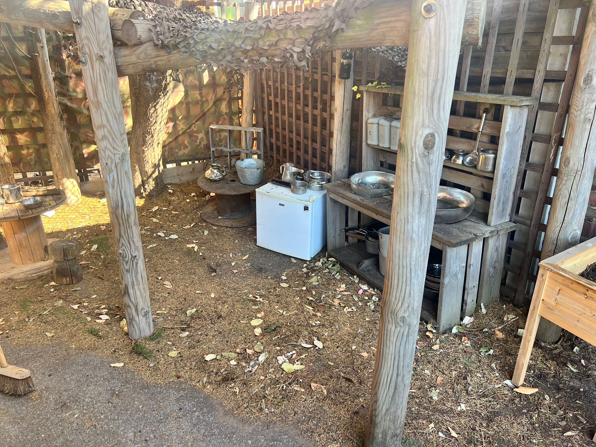 Wooden shelving and a small white mini-fridge hold metal bowls, pots and utensils, arranged under a lattice-roofed, camouflaged canopy in a shaded outdoor play/kitchen area with leaf-strewn ground.