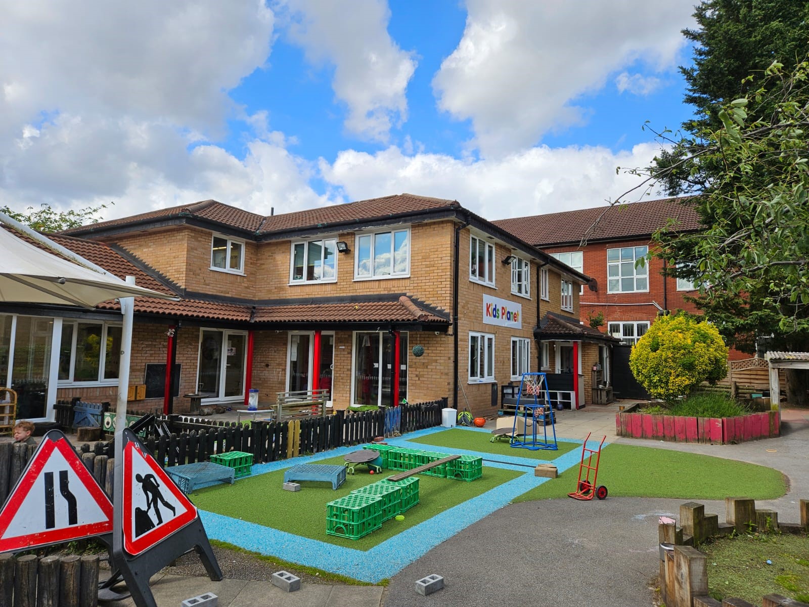Two-story daycare building (sign: 'Kids Planet') overlooks an outdoor play area with toy crates, a small basketball hoop, traffic warning signs, and artificial turf under a cloudy blue sky.