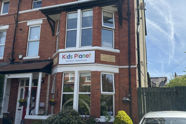 Red-brick building with a "Kids Planet" sign (text: "Kids Planet - Day Nursery - Meithrinfa Dydd") mounted above a bay window, flanked by hanging baskets on a sunny residential street.