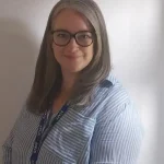 Woman with shoulder-length gray hair and glasses smiles at camera, standing before a plain light wall; she wears a blue-striped shirt and a lanyard labeled 
