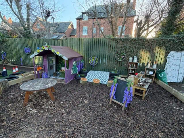 Purple wooden playhouse sits decorated with floral garlands; surrounded by a round wooden table, chalkboard, baskets and toy kitchen; set on woodchip ground inside a fenced suburban backyard.