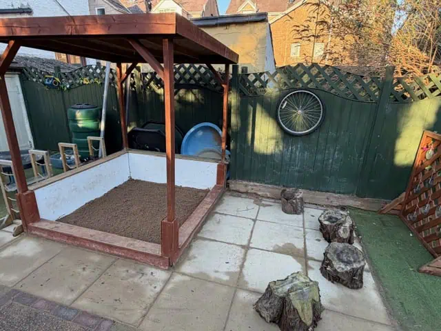 Wooden sandbox sits filled with gravel under a small pergola; several tree-stump seats line the paved patio; a green fence with a mounted bicycle wheel and garden tubs surrounds the backyard.