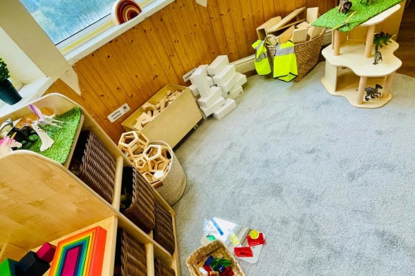 A carpeted children's play area: wooden shelves and baskets hold colorful blocks and toys, small high-visibility vests hang nearby, and natural light enters through a window over wood-paneled walls.
