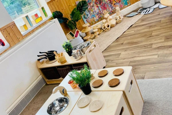 Wooden play kitchen sits stocked with metal sink, toy stove burners, pots, pans and potted plants, arranged on modular units in a sunlit playroom with wooden floor, baskets and art.