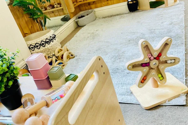 Wooden star-shaped bead maze holds colorful beads in channels, sitting on a light-gray rug in a sunlit children's playroom with wooden toys, pastel stacking boxes, a plant and woven baskets.