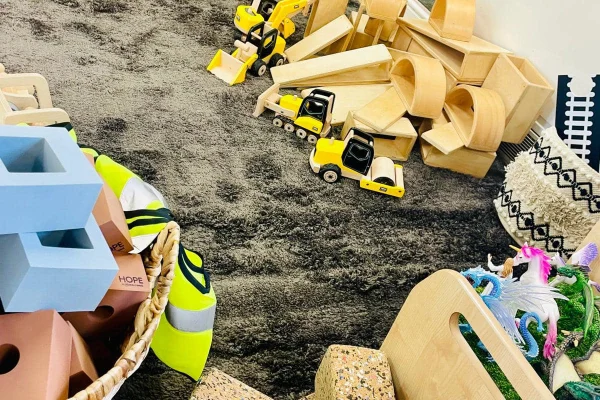 Yellow toy construction vehicles line up beside wooden ramps and blocks, surrounded by pastel blocks and a basket with neon safety vests labeled "HOPE" on a dark gray carpet.
