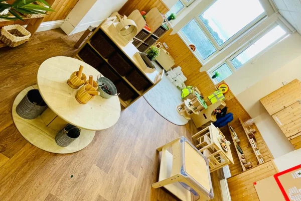 Low round wooden table holds small baskets, bowls, and wooden toys, arranged on laminate floor amid child-sized tables, shelves, a play kitchen, and windows in a bright, wood-paneled preschool classroom.
