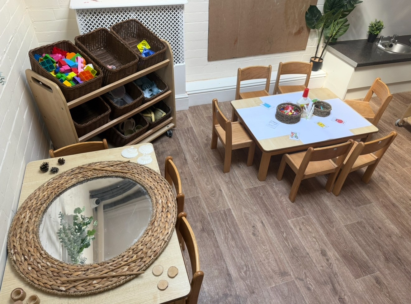Low wooden tables and chairs — hold baskets of craft supplies and a braided mirror — arranged for small-group play in a corner preschool classroom with storage shelves, sink and wood-look floor.