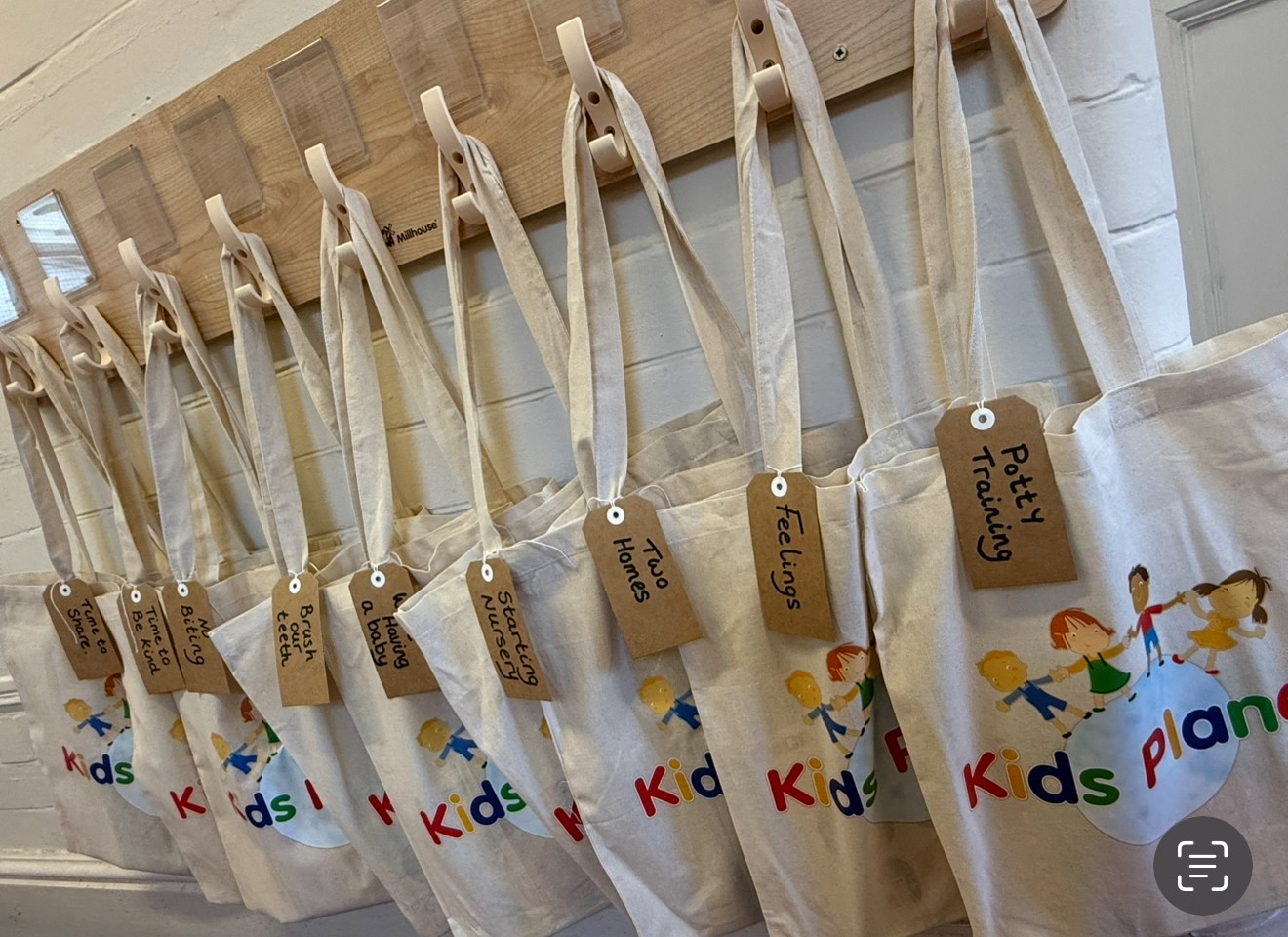 Row of canvas tote bags hanging from wooden hooks in a tiled hallway. Kraft tags read: "Time to share?", "Be kind", "Brush teeth", "Leaving a baby", "Starting nursery", "Two Homes", "Feelings", "Potty Training". Bags show "Kids Planet" logo.