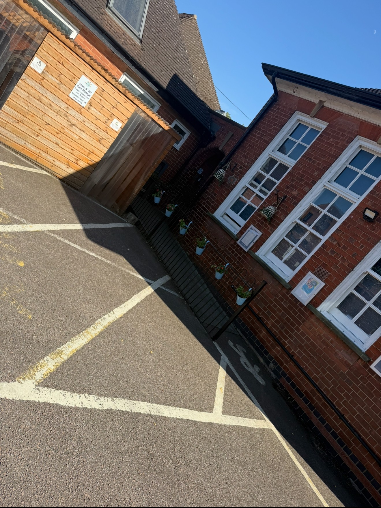 Red-brick building displays hanging potted plants and windows; adjoins a wooden shed and marked parking bays on asphalt under a clear blue sky. Small signs visible but illegible.