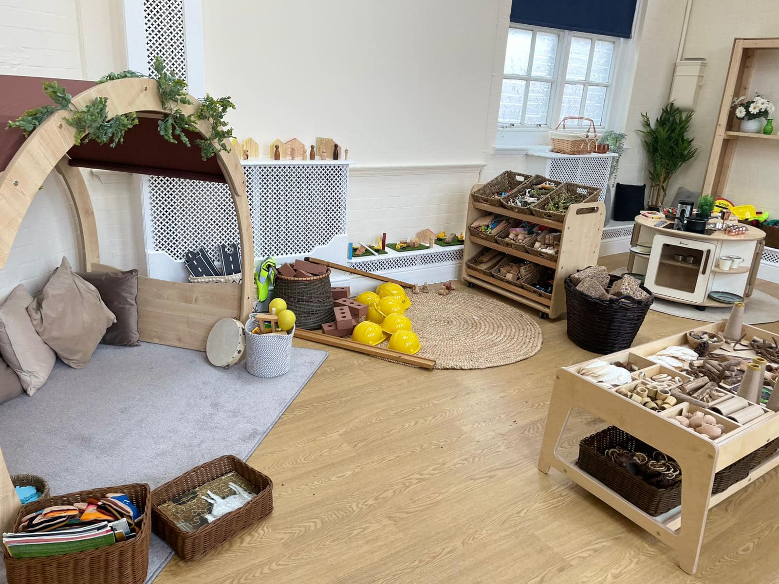 Wooden play arch shelters cushions and a tambourine, surrounded by baskets of natural toys; yellow toy construction helmets spill onto a round rug in a preschool room with shelves.