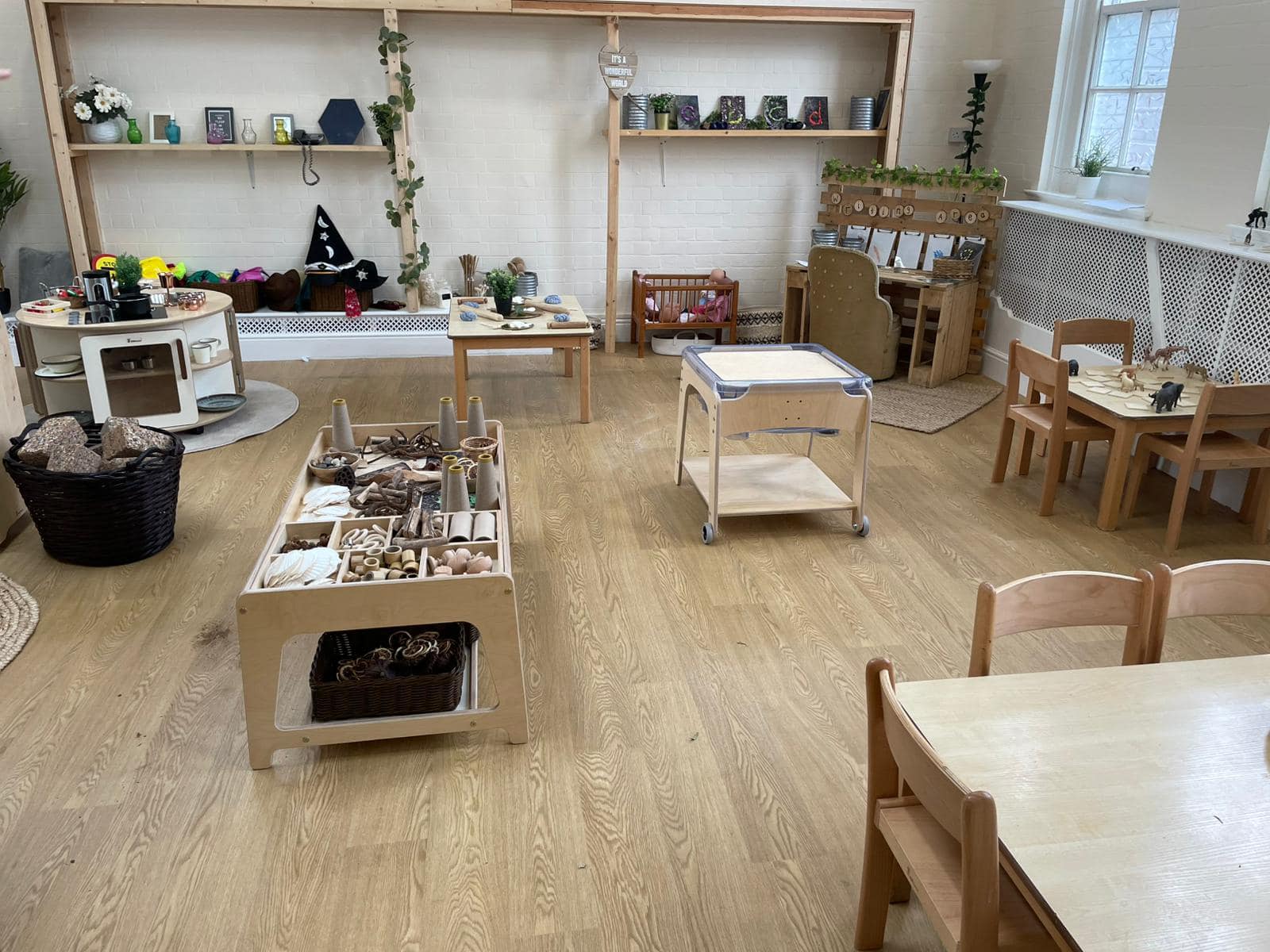 Children's wooden activity tables and low shelves hold toys and natural materials, arranged for play across a sunlit classroom with small chairs, a toy kitchen, and wooden flooring. Text: "IT'S A WONDERFUL WORLD"