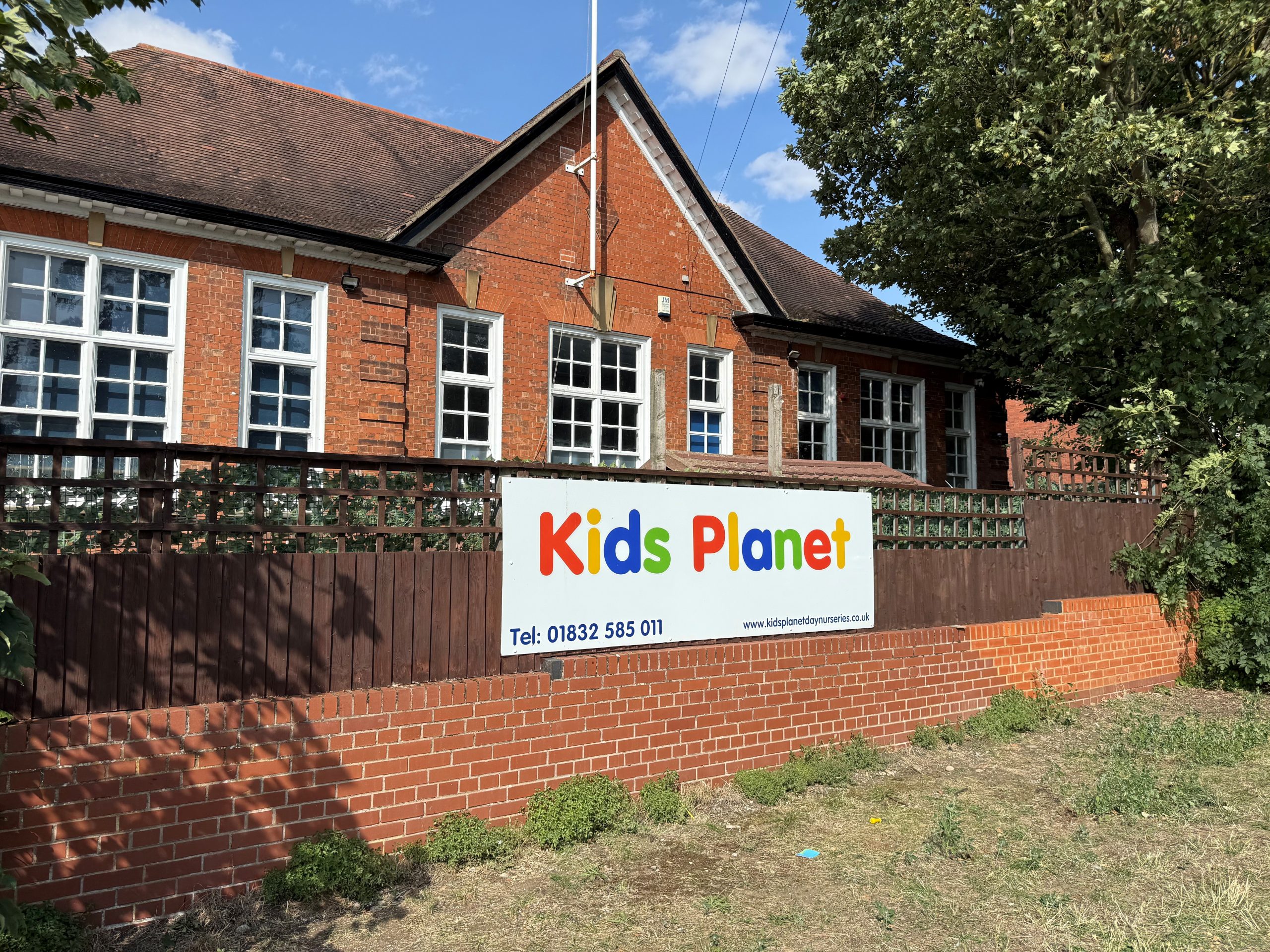 A sign reading "Kids Planet" (Tel: 01832 585 011, www.kidsplanetdaynurseries.co.uk) is mounted on a fence in front of a red-brick building with white-paned windows, trees, and a blue sky.
