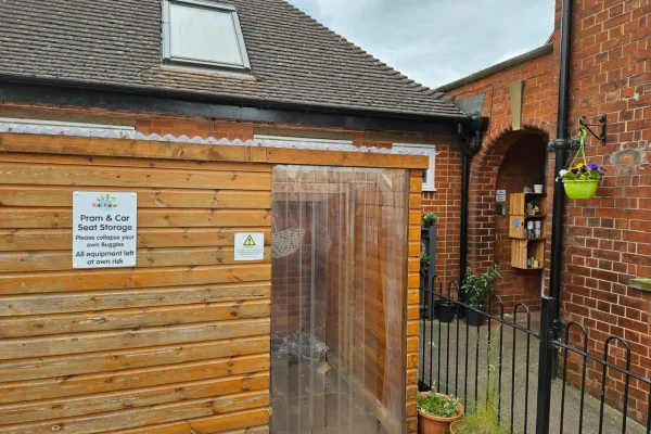 Wooden storage shed with clear plastic strip door, housing prams/car seats; small brick courtyard with arched entry, potted plants and a low metal gate.