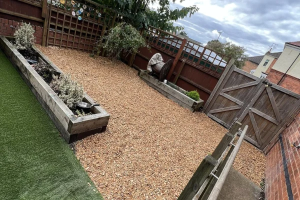 Gravel-covered backyard occupies center, featuring two raised wooden planters and a covered lawn ornament; enclosed by wooden fencing and a gate beside brick walls, artificial turf at left, overcast sky.