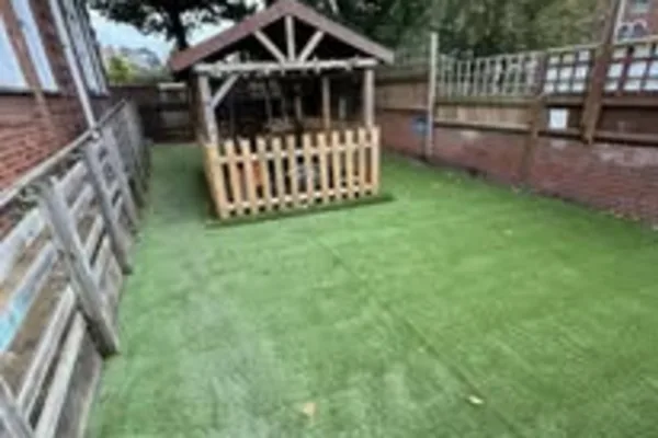 Small wooden playhouse sits within a picket-fenced platform, standing on artificial turf in a narrow, fenced backyard bordered by brick walls and wooden panels.