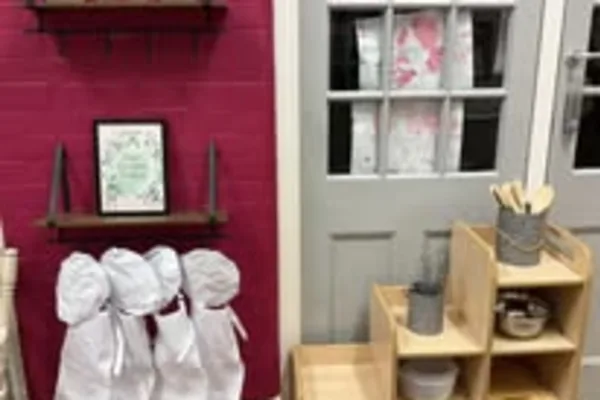 Four small chairs draped in white cloth stand against a red brick wall; a framed print and wooden shelving with kitchenware sit beside a gray door featuring divided window panes.