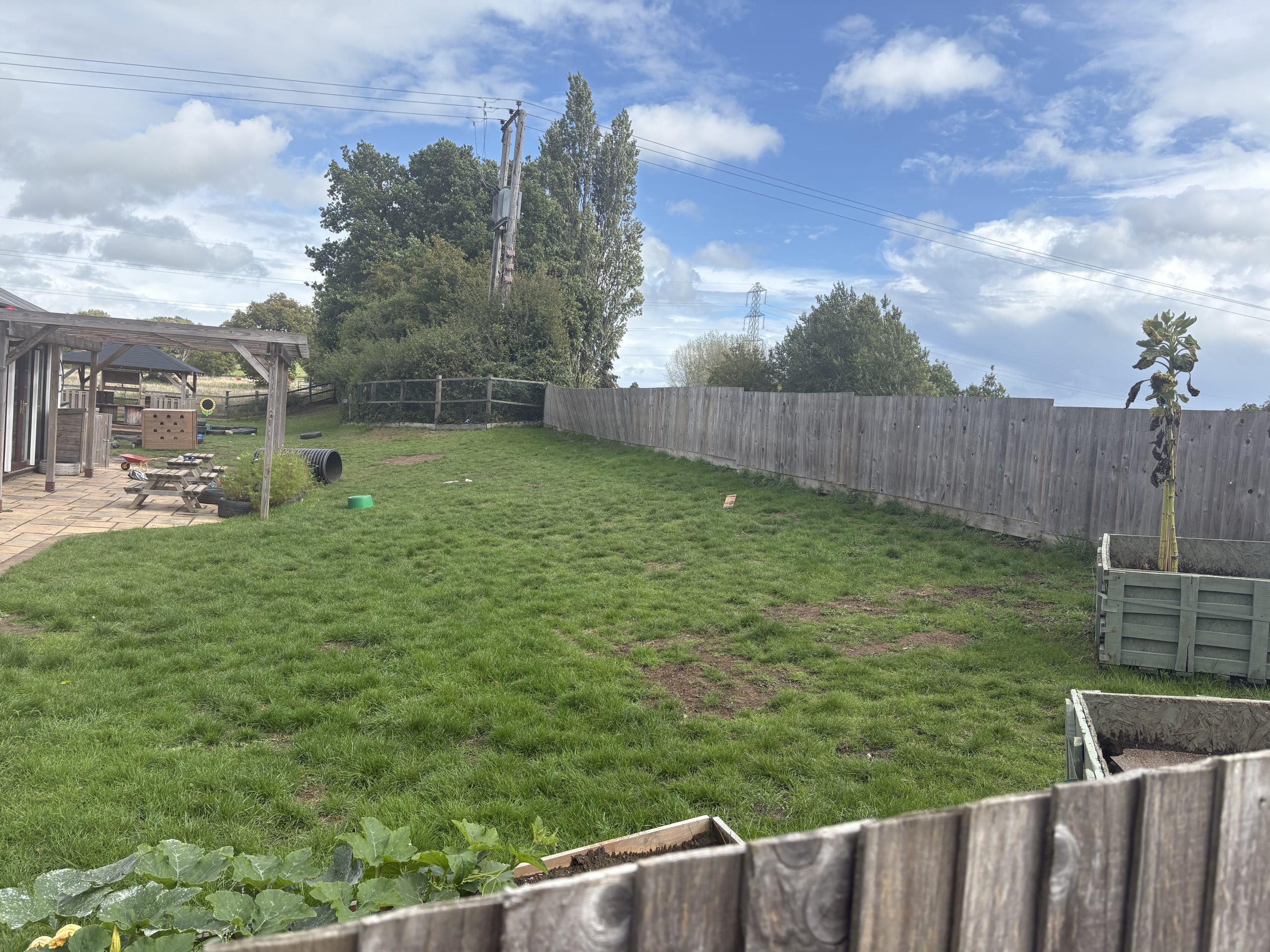 A grassy backyard slopes upward, bordered by a tall wooden fence; a pergola and picnic table sit left with play items, planter boxes and trees under a partly cloudy sky.