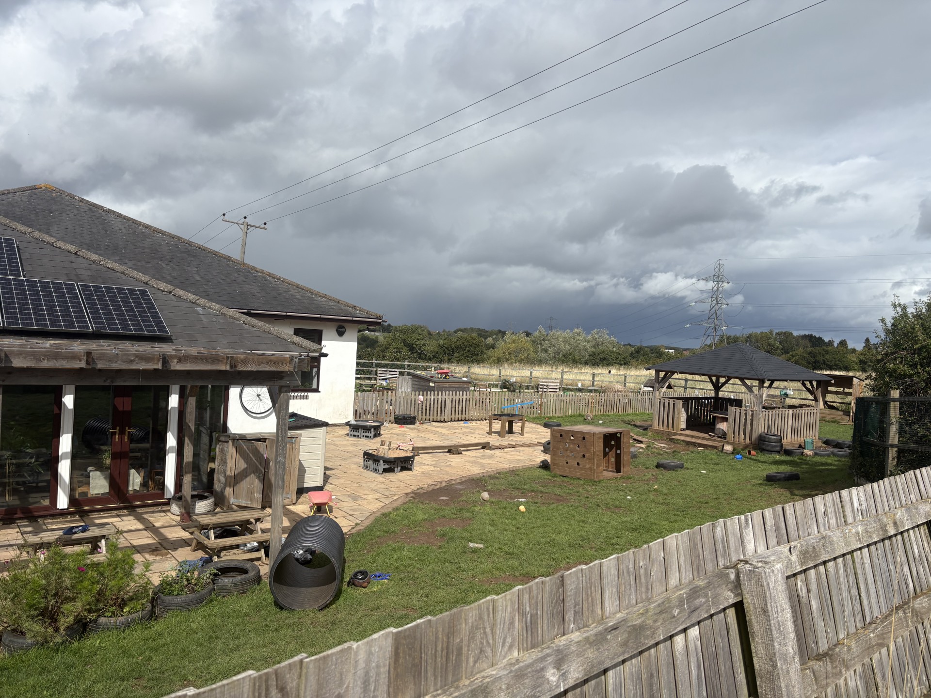 Single-story building with solar panels overlooks a fenced playground; children’s play equipment—wooden gazebo, tire tunnel, climbing box, scattered toys—occupies a grassy yard under dark, cloudy sky.
