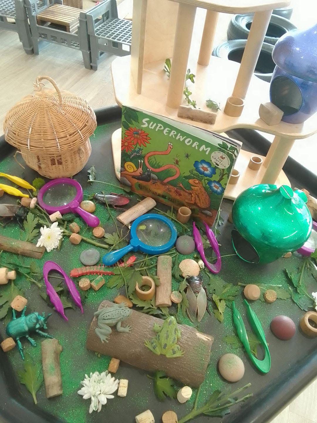 A children's book titled SUPERWORM stands upright among plastic magnifiers, toy insects, corks, logs and leaves in a green-glitter sensory play tray on a classroom floor with wooden shelves.