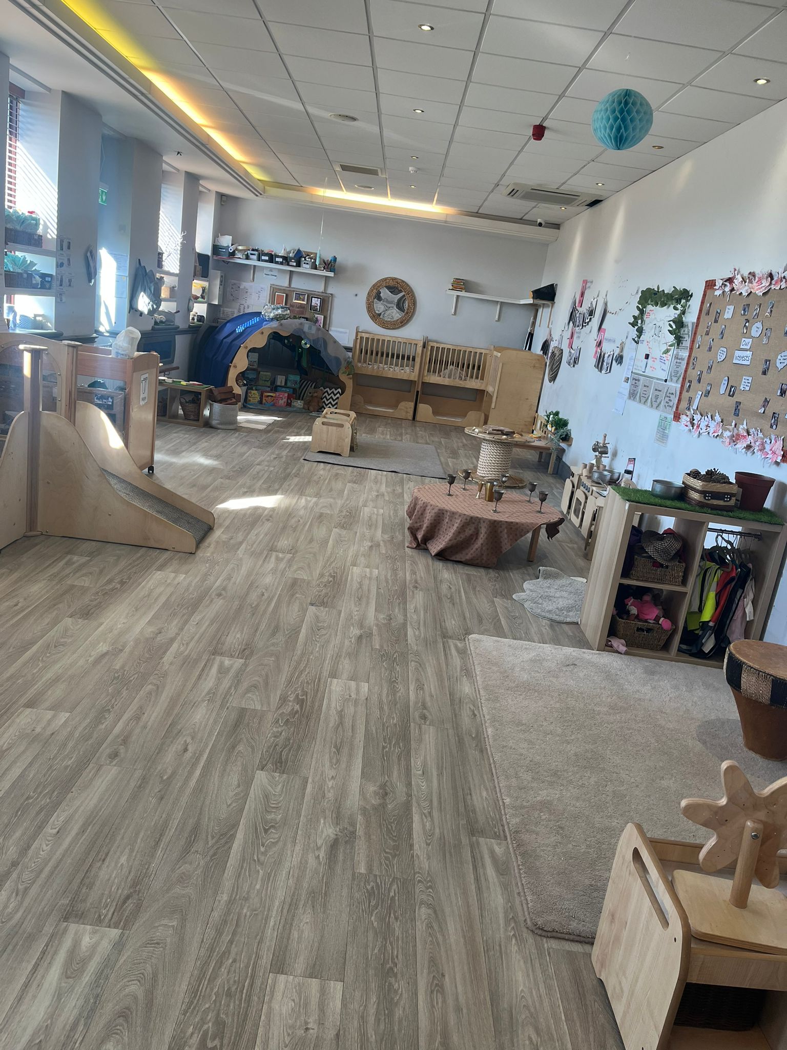 Children's wooden play furniture sits arranged for use, surrounded by rugs, shelves, and a reading tunnel inside a bright, spacious classroom with wood-look flooring and wall displays.