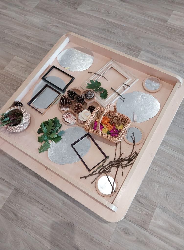 Wooden tray holds arranged natural materials—pinecones, oak leaves, shells, sticks, small mirrors, empty picture frames and two baskets of petals—placed on a light laminate floor.
