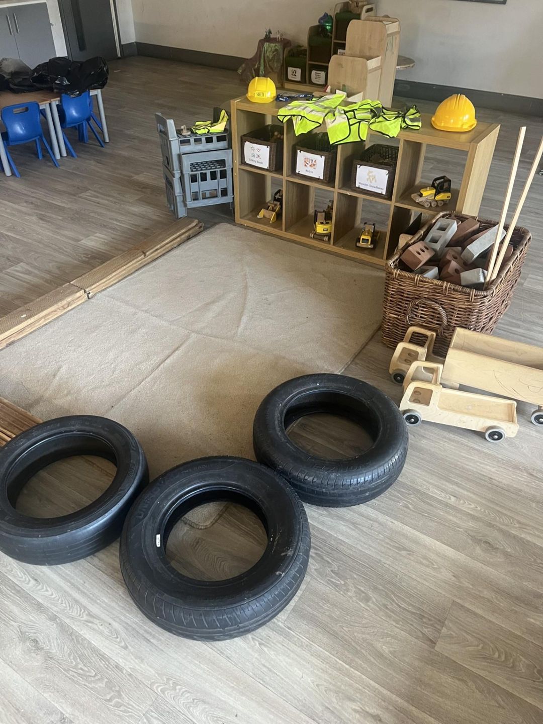 Three black tires lie on a floor in an indoor play area beside a rug, toy trucks, a basket of blocks, and cubby shelves holding safety vests and hard hats.