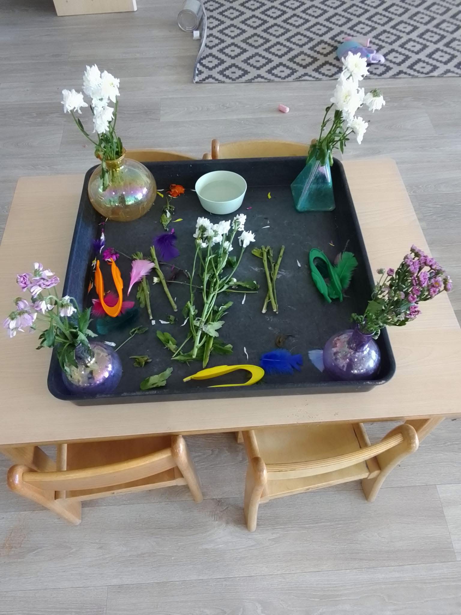 Black tray holds glass vases with white and purple flowers, loose stems, colorful feathers and plastic scoops; a pale bowl sits center on a low wooden table in a light-floored room.
