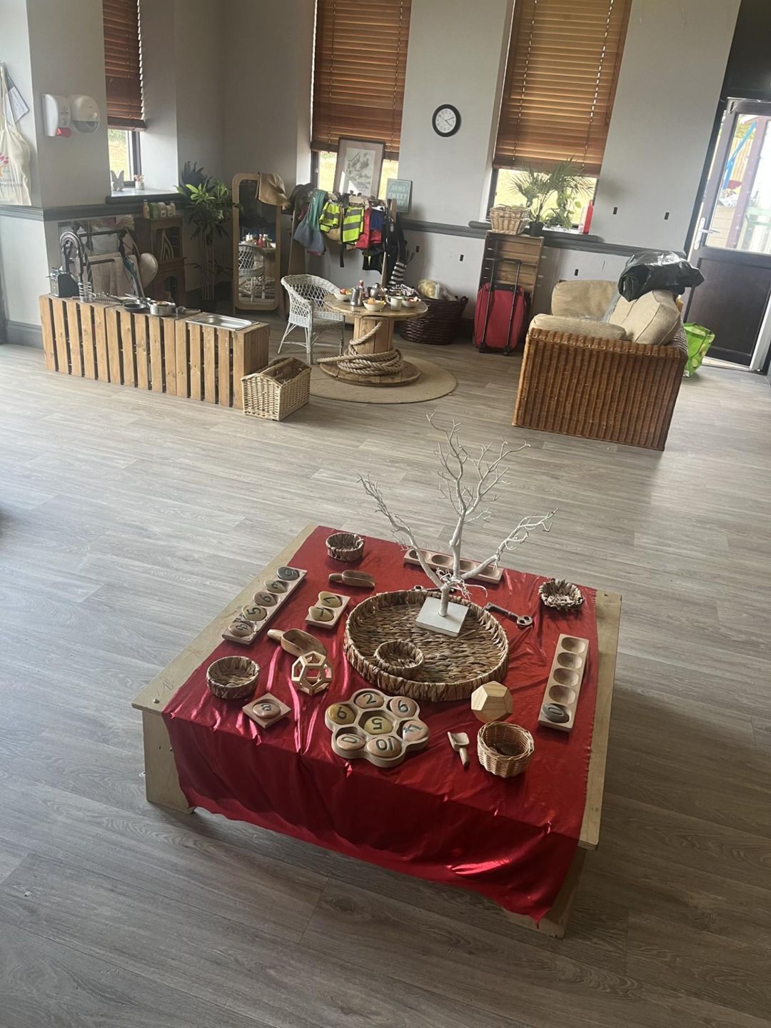 Low wooden play table draped with red fabric displaying wooden toys, baskets and a white twig tree, in a spacious light-filled playroom with wicker seating, coat rack and windows.
