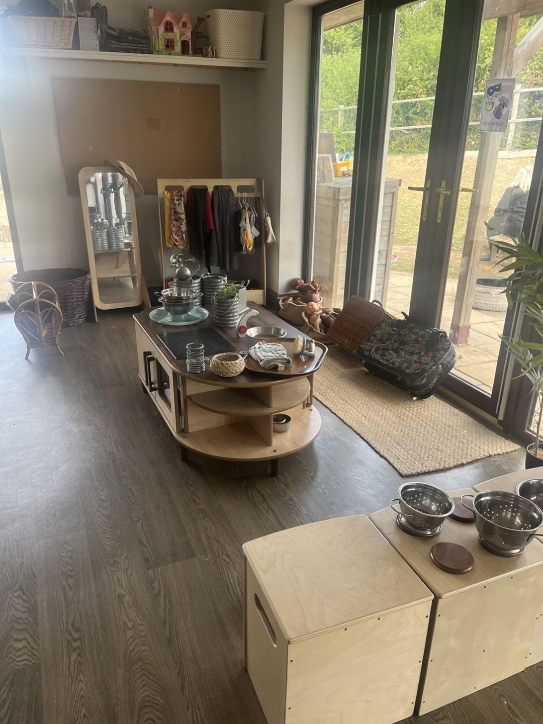 Low play table holds toy pots, bowls and utensils; nearby stools with metal colanders, a mirror and coat rack with children's clothes, glass doors opening to a grassy patio. Text: "Dressing up".