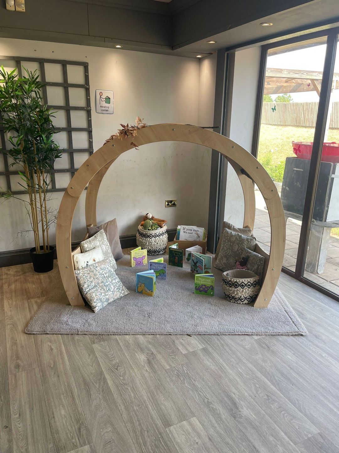 Curved wooden arch shelters cushions, baskets and children's board books for reading; placed on a rug beside floor-to-ceiling glass doors and a potted plant in a bright indoor corner.