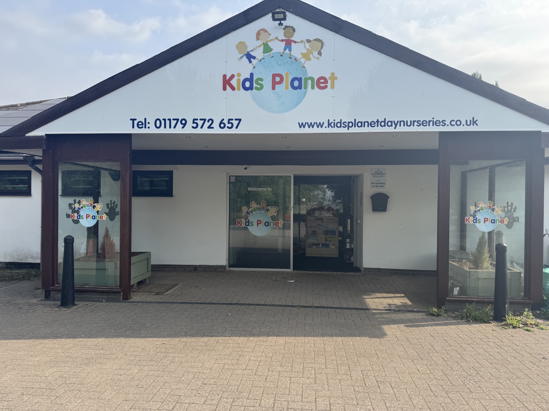 Large "Kids Planet" sign and logo mark a nursery entrance; it displays contact details above glass doors, flanked by planters and bollards on a paved forecourt.
