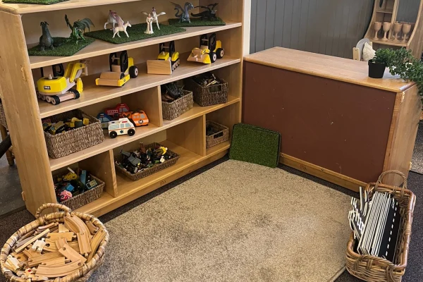 Wooden shelving displays toy vehicles, animal figures and woven baskets; nearby baskets hold wooden tracks and folders, against a carpeted playroom corner with a small counter and green play mat.