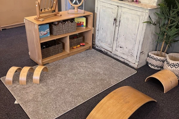 Curved wooden balance boards rest on and beside a gray rug; low wooden shelf holds woven baskets and children's toys, positioned against a distressed white cabinet in a playroom corner.