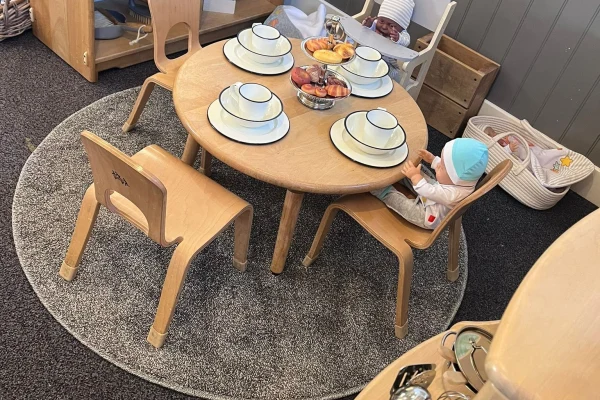 Small round wooden children's table set for a tea party with four stacked white bowls and a toy-fruit centerpiece; two dolls sit in chairs on a circular gray rug in a play area.