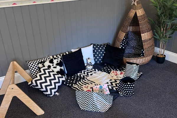 Cluster of black-and-white patterned cushions and fabric bins arranged on a carpet, forming a cozy reading corner next to a wicker tepee and wooden climbing frame in a gray-paneled room.
